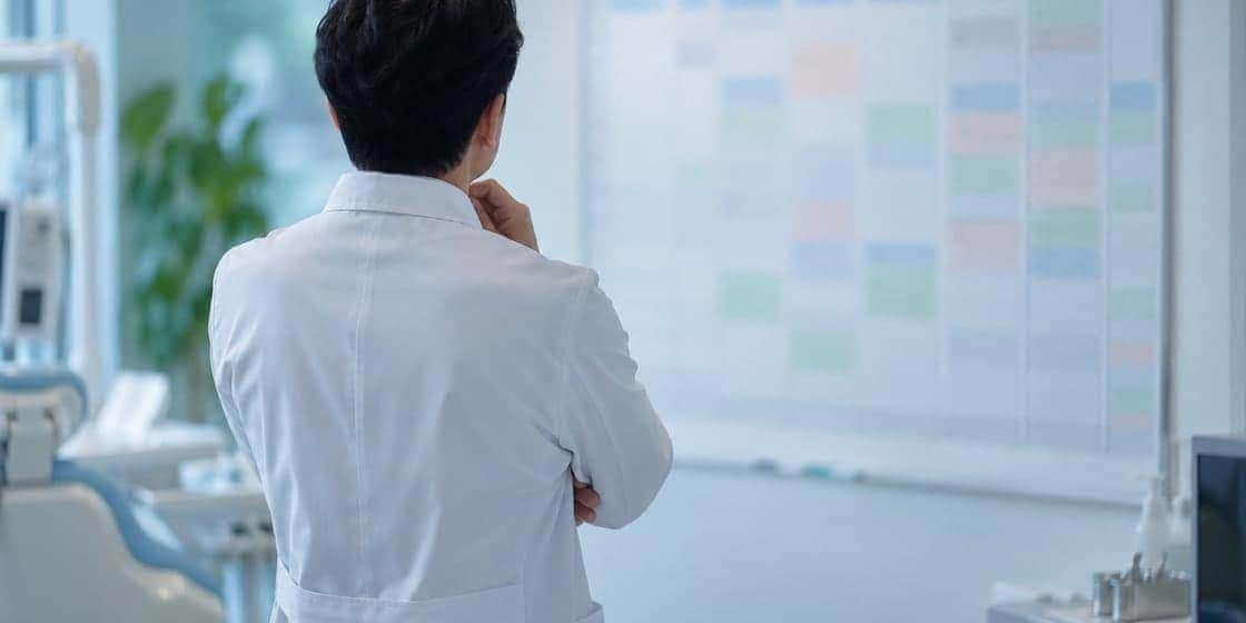 週間予定表の前に立つ白衣姿の歯科医師の後ろ姿